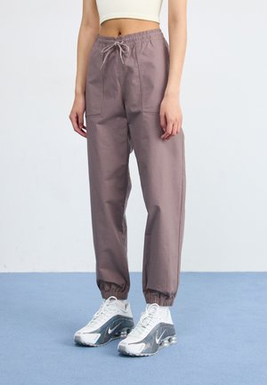 Une personne se tient debout dans un pantalon de jogging violet clair et des baskets blanches avec des détails réfléchissants, devant un fond uni et un sol bleu doux.