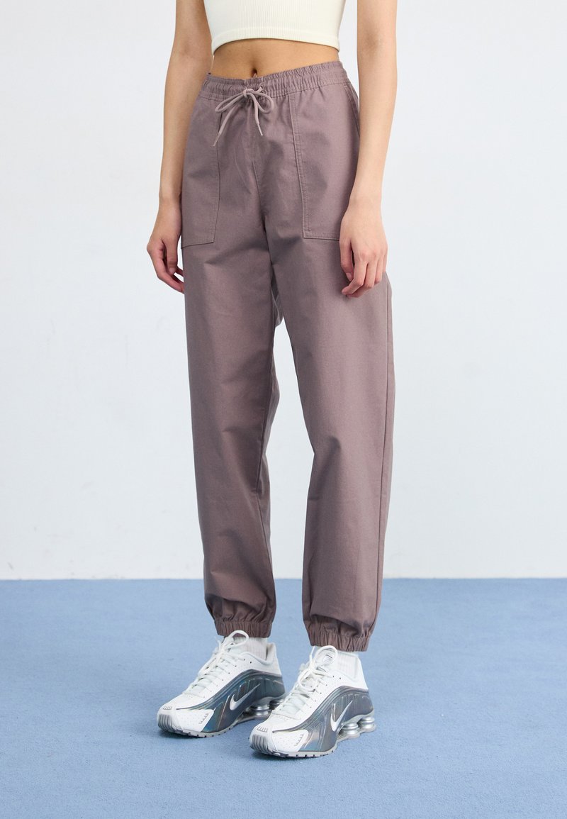 A person stands in light purple joggers and white sneakers with reflective details, against a plain background and soft blue flooring.