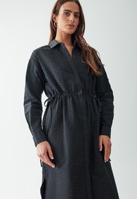 Abito camicia nero realizzato in tessuto texturizzato, con chiusura a bottoni sul davanti e vita con cordoncino; presenta maniche lunghe e spacchi laterali per facilitare il movimento.