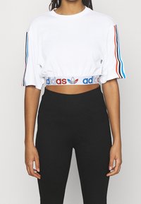 Bílý bavlněný krátký tričko s krátkými rukávy, s modrými, červenými a černými pruhy na rukávech a elastickým páskem s logem v pase.