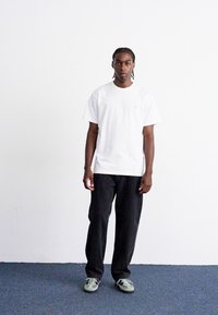 Weißes Baumwoll-T-Shirt mit kurzen Ärmeln, rundem Ausschnitt und kleinem goldenen Logo, kombiniert mit dunklen, lockeren Hosen und grauen Sneakers.