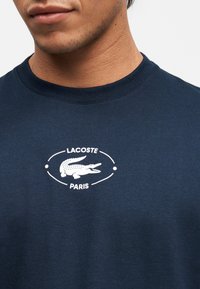 Άνδρας που φοράει μπλε ναυτικό t-shirt με στρογγυλή λαιμόκοψη και λευκό λογότυπο Lacoste Paris με το έμβλημα κροκόδειλου στο στήθος.