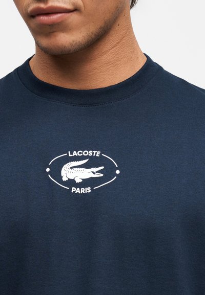 Мъж, облечен в тъмносин тениска с бяла емблема на Lacoste Paris и крокодил на гърдите.