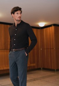 Donkerbruine gebreide poloshirt met een kraag, voorzien van witte knopen, gecombineerd met grijze gestructureerde broek en een geweven bruine riem. Houten achtergrond.