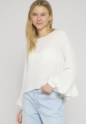 Junge Frau mit langen blonden Haaren, die eine lockere weiße Bluse mit langen Ärmeln und hellblaue, hoch taillierte Jeans trägt, steht mit der Hand in der Tasche.