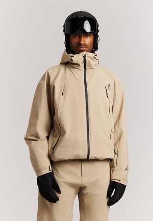 Man met een beige skipak en broek, zwarte skihandschoenen en een zwarte skihelm met bril tegen een effen achtergrond.