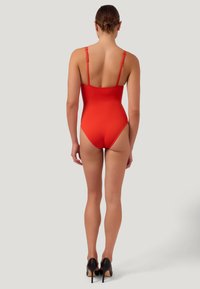 Röd baddräkt i ett stycke med justerbara axelband, slät textur och hög skärning vid benöppningarna, parat med svarta högklackade skor.