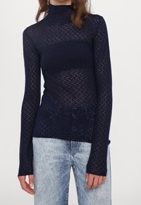 Top blu navy a maniche lunghe in maglia con collo alto, caratterizzato da un design traforato testurizzato e silhouette aderente. Indossato sopra jeans azzurri.