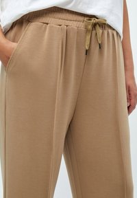 Beige elastische broek met een koordsluiting in de taille. Bevat zijzakken en heeft een gladde textuur, ontworpen voor comfort en bewegingsvrijheid.