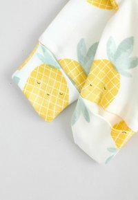 Bomullstyg med vit bakgrund har ett mönster av gula ananaser med gröna blad. Ärmarna är rundade och mjuka vid beröring.