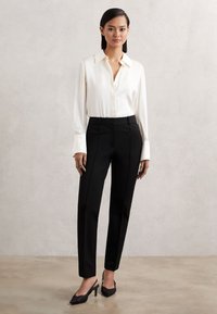 Kvinde stående op ad en lys væg iført en hvid bluse med lange ærmer, sorte skræddersyede bukser og sorte slingback sko med hæl.