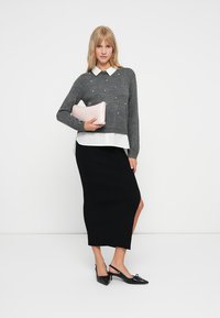 Suéter de punto gris con perlas, camisa blanca con cuello por debajo, falda midi negra con canalé, sosteniendo un bolso de mano rosa claro y usando unas bailarinas negras con tacón tipo slingback.