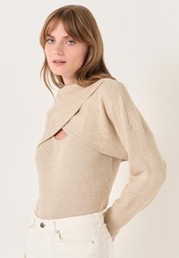Pullover beige a costine con un design incrociato unico e un'apertura circolare sul collo. Tessuto morbido e maniche lunghe.