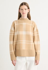 Sweater de lã bege com um decote alto canelado, corte oversized e padrão de quadrados brancos. Textura suave e punhos canelados nos pulsos.