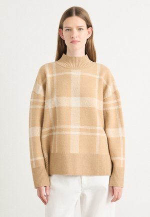 Beige wollen sweater met een hoge ribbelkraag, oversized pasvorm en een wit ruitpatroon. Zachte textuur en ribbelmanchetten bij de polsen.
