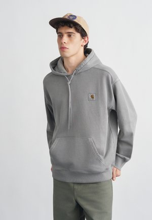 Młody mężczyzna w szarej bluzie Carhartt z kapturem i beżowej czapce z daszkiem, stojący na jasnym, jednolitym tle, lekko zwrócony w lewo.