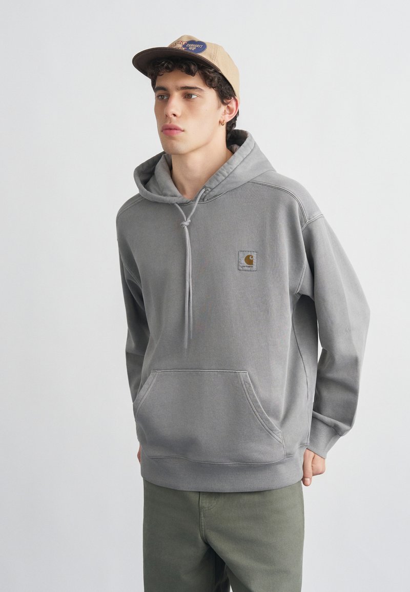 Νεαρός άνδρας φορώντας γκρι φούτερ Carhartt με κουκούλα και μπεζ καπέλο μπέιζμπολ, στέκεται μπροστά σε απλό ανοιχτό φόντο, στραμμένος ελαφρώς προς τα αριστερά.