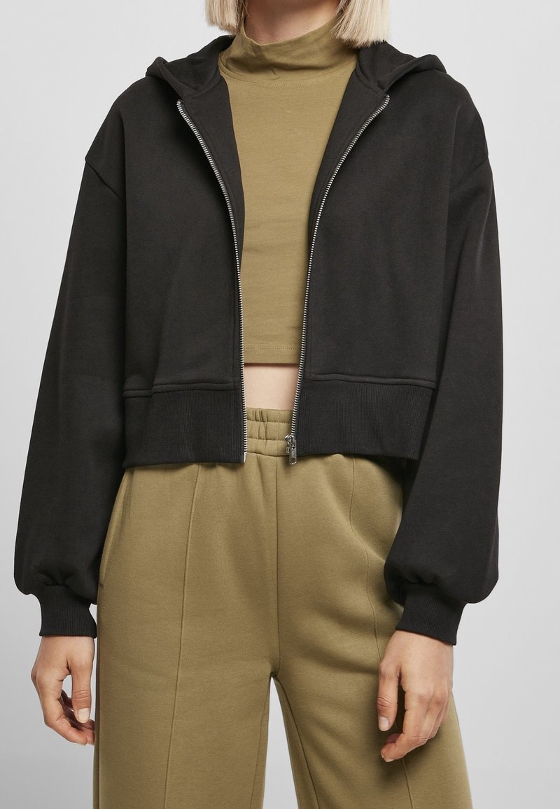 Woman wearing black cropped zip-up hoodie over olive green high-neck crop top and matching high-waisted pants, standing against gray background.