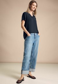 Blusa nera a maniche corte con scollo a V abbinata a jeans larghi in denim blu chiaro, con risvolti, e scarpe basse in rete nere. Tessuto dalla superficie liscia.