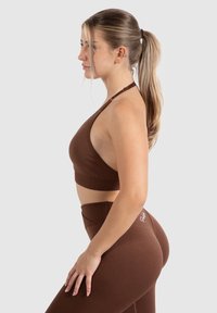 Conjunto de entrenamiento marrón que incluye un sujetador deportivo de cuello halter y leggings de tiro alto. Textura suave, diseño minimalista y silueta ajustada.