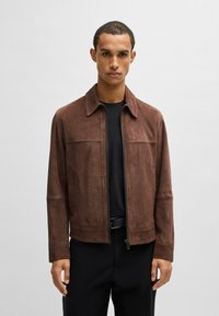 Giacca in suede marrone con zip frontale, colletto aperto, due tasche anteriori e texture liscia, indossata sopra una maglietta nera.