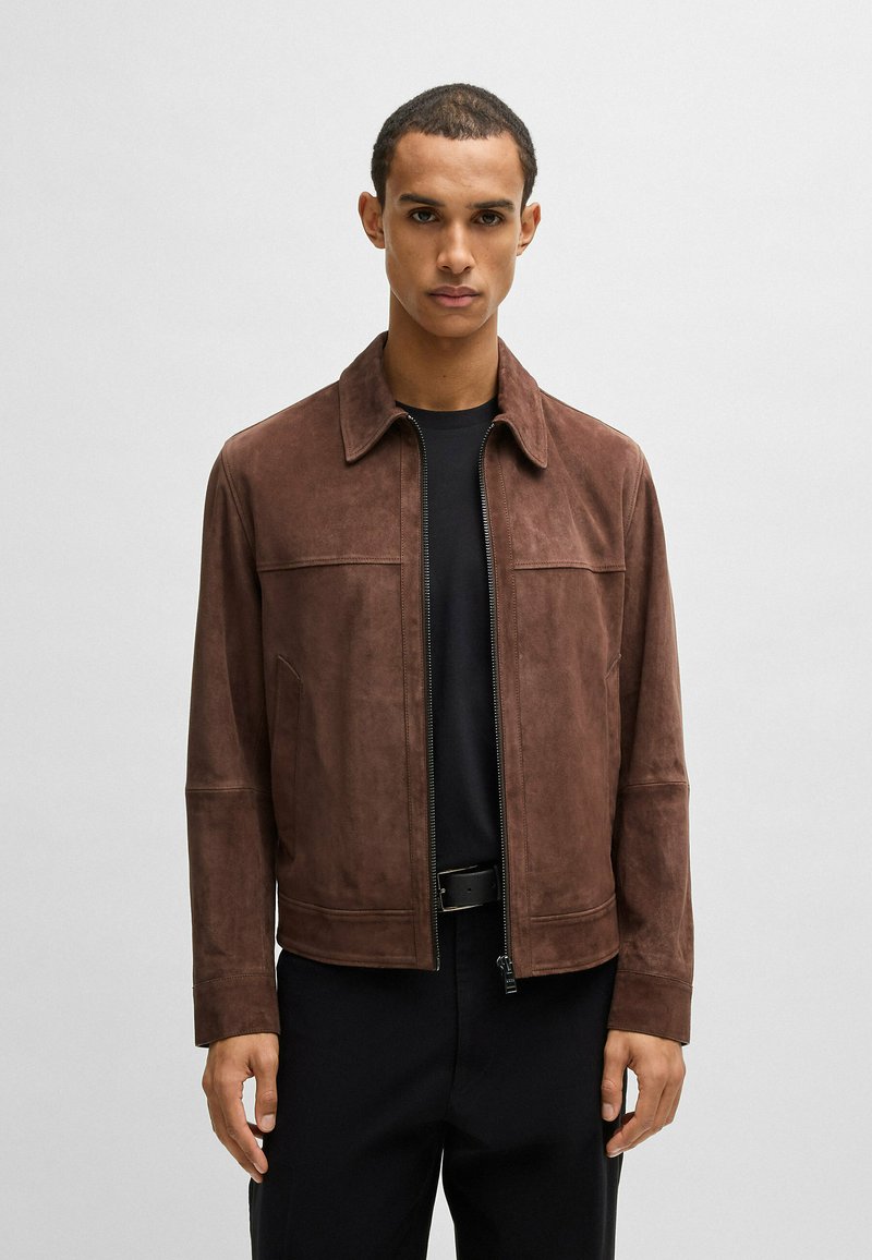 Giacca in suede marrone con zip frontale, colletto aperto, due tasche anteriori e texture liscia, indossata sopra una maglietta nera.