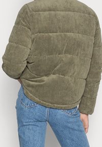 Chaqueta puffer de pana en verde oliva con diseño acolchado. Dobladillo redondeado y mangas largas, combinada con jeans de mezclilla azul claro.