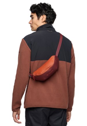 Verstellbare Umhängetasche aus rot-orangefarbenem Stoff mit schlanker Form, ausgestattet mit einem Reißverschluss und Logo-Details, über der Schulter getragen.