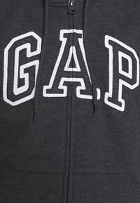 Donkergrijze zip-up hoodie met grote witte geborduurde "G.A.P" letters op de voorkant, met een textuur stof en een hoodie met trekkoord.