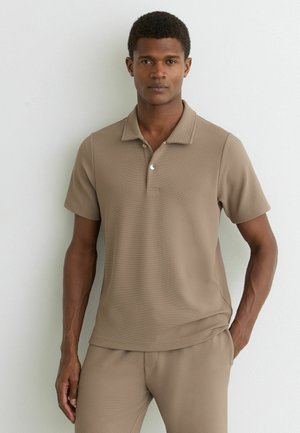 Muž stojaci opretý o bielu stenu, oblečený v béžovej polokošeli s krátkym rukávom a zladených nohaviciach, s jednou rukou vrecku.