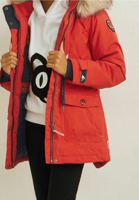 Czerwona kurtka puchowa z kapturem wyłożonym futrem, zamek błyskawiczny i zatrzaski, oraz kieszenie po bokach; zestawiona z białym bluzą z kapturem z czarną grafiką.
