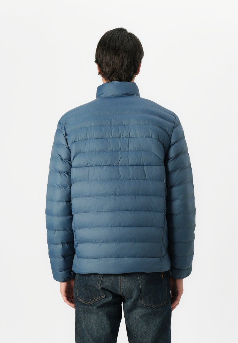 Chaqueta acolchada azul con secciones acolchadas horizontales, cuello alto y puños elásticos, combinada con jeans de mezclilla oscura. Textura suave.
