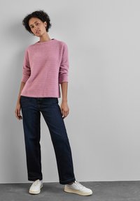 Rosa flauschiger Pullover mit Rundhalsausschnitt, kombiniert mit dunkelblauen geraden Jeans und weißen Sneakers, vor einem neutralen Hintergrund.