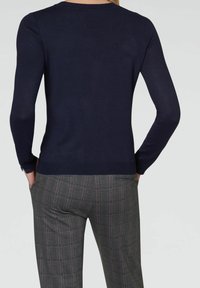 Femme portant un pull bleu marine à manches longues et un pantalon à carreaux gris, les mains dans les poches, vue de dos et de la taille jusqu'en haut.