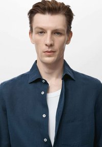 Námornícka modrá ľanová košeľa s golierom na gombíky, s vreckom na hrudi a bielymi gombíkmi. Nosená cez bielu tielko. Jednoduchý, textúrovaný dizajn.