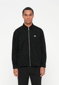 Juoda zip-up striukė su tekstūriniu deimantiniais raštais, smailiu apykakle ir mažuoju logotipo užnneriu ant kairiojo krūtinės. Ilgos rankovės su rankogaliais.