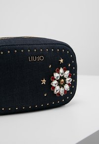 Borsa cosmetica in denim blu scuro con borchie dorate, caratterizzata da un'applicazione floreale in strass e il logo del marchio "LIUJO" in oro.