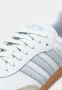 Baltie Adidas Samba sporta apavi ar gaiši zili svītraini un logo, baltas auklas un gumijas zole, attēlots tuvplānā sānskatā.