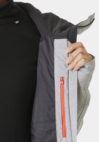 Šedá bunda s oranžovým zipem, ukazující vnitřní vrstvu černé tkaniny. Má hladkou texturu a strukturovaný design pro teplo.