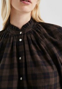 Bruine geruite blouse met een hoge hals, gathered stof en drie witte knopen aan de voorkant. Zachte textuur met zichtbaar patroon.
