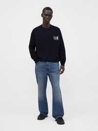 Homme portant un sweat-shirt noir avec un logo sur la poitrine, un jean bleu ample et des baskets noires, debout devant un fond uni.