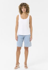 Weißes ärmelloses Top, hellblaue Denim-Shorts und beige Riemchen-Heels. Das Model steht vor einem schlichten Hintergrund.