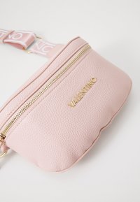 Rozā ādas jostas soma ar raupju tekstūru, zelta rāvējslēdzējiem un logotipu. Iezīmēta ar izdrukātu regulējamu siksnu rozā un baltās krāsās.