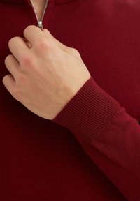 Sweatshirt zippé rouge avec des poignets côtelés, montrant une main tenant la fermeture éclair. Le tissu semble doux, avec une texture lisse.