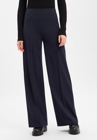 Marineblå vide bukser laget av mykt stoff med en jevn tekstur, med høy midje og avslappet passform. Brukes med svarte sko.