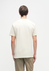 Ανοιχτό μπεζ βαμβακερό μπλουζάκι με κοντά μανίκια και στρογγυλή λαιμόκοψη. Έχει λεία υφή και χαλαρή εφαρμογή. Δεν υπάρχουν ορατά γραφικά ή σχέδια.