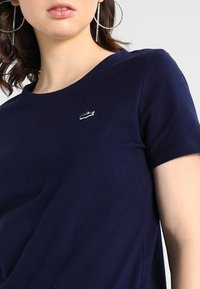 Námořnická modrá bavlněná tričko s kulatým výstřihem, krátkými rukávy a malým vyšívaným logem na hrudi. Hladká textura.