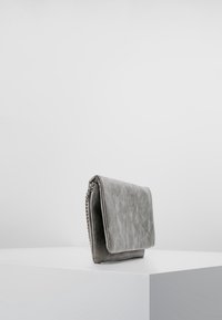 Sac à main en simili cuir gris, de forme rectangulaire, avec un rabat replié et un détail en chaîne sur le côté. Texture lisse et design minimaliste.