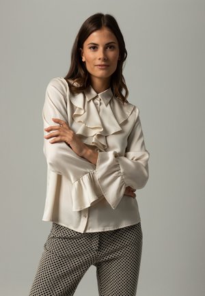 Cream blouse with ruffled collar and sleeves, featuring a smooth texture and button closure, paired with patterned dark trousers.