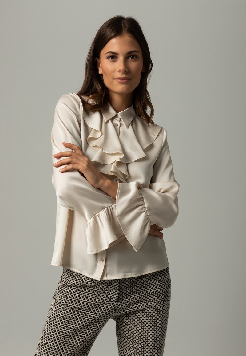 Cream blouse with ruffled collar and sleeves, featuring a smooth texture and button closure, paired with patterned dark trousers.